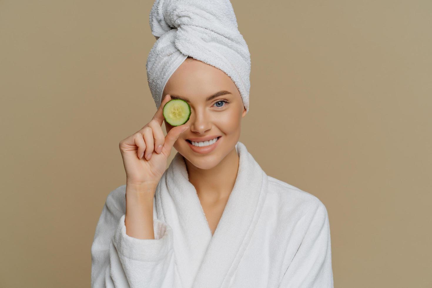Horizontal shot of pleased refreshed woman covers eye with slice of fresh cucumber cares about skin uses organic products smiles pleasantly dressed in bath dressing gown after taking shower. photo