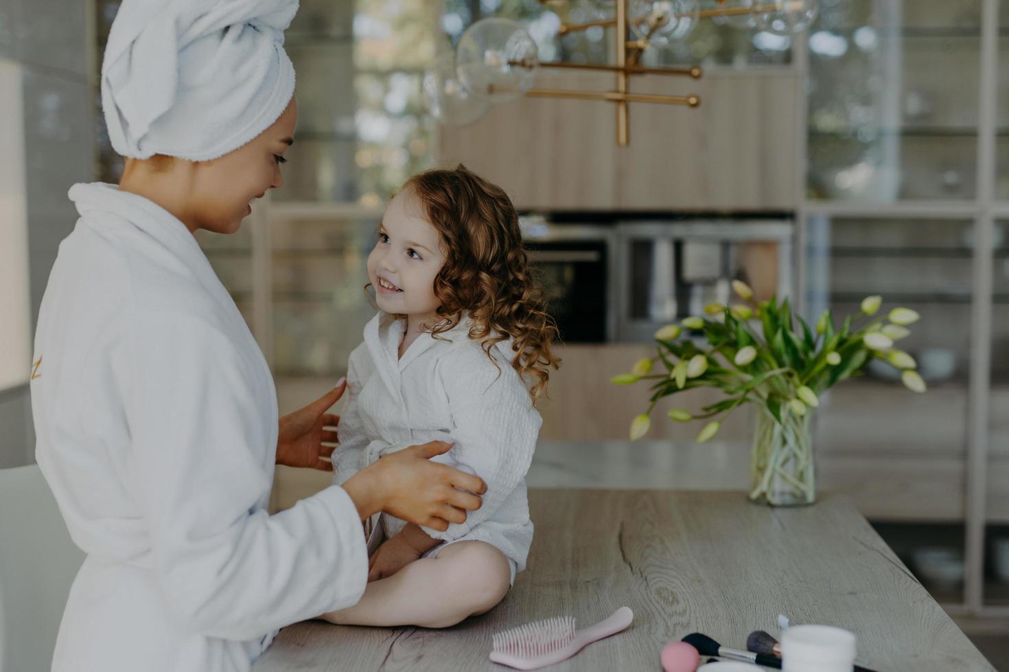 madre e hija en batas de baño blancas tienen una agradable conversación entre ellas y van a someterse a procedimientos de belleza después de ducharse. pequeña niña adorable con el pelo rizado mira a mamá en casa. foto