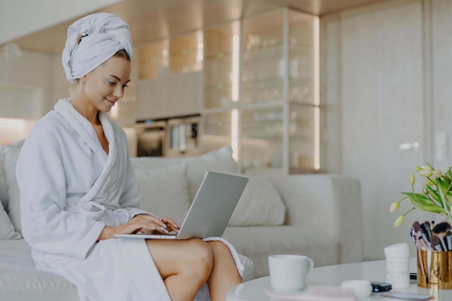 Una foto horizontal de una mujer hermosa satisfecha vestida con un albornoz suave blanco trabaja en una computadora portátil navegando por Internet posa cerca de la mesa con bebidas y productos cosméticos sentados en un cómodo sofá