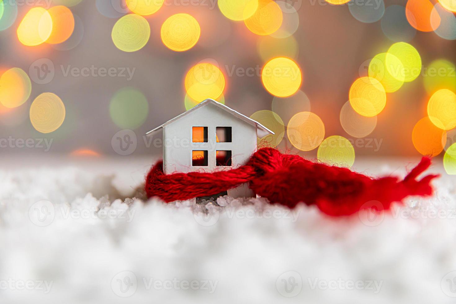 Abstract Advent Christmas Background. Toy model house with knitted red scarf on snow with defocused garland lights background. Christmas with family at home concept. Christmas winter composition. photo