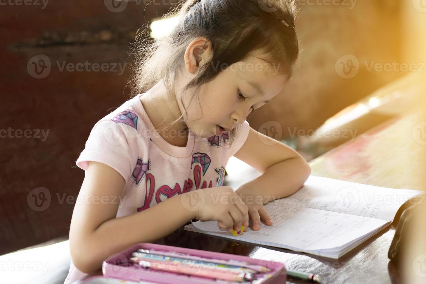 chica asiática sentada en un escritorio en casa escribiendo un libro. educación en línea. nueva normalidad educativa. conceptos de lectura y escritura de los niños. educación desde casa. borrar el error tipográfico foto