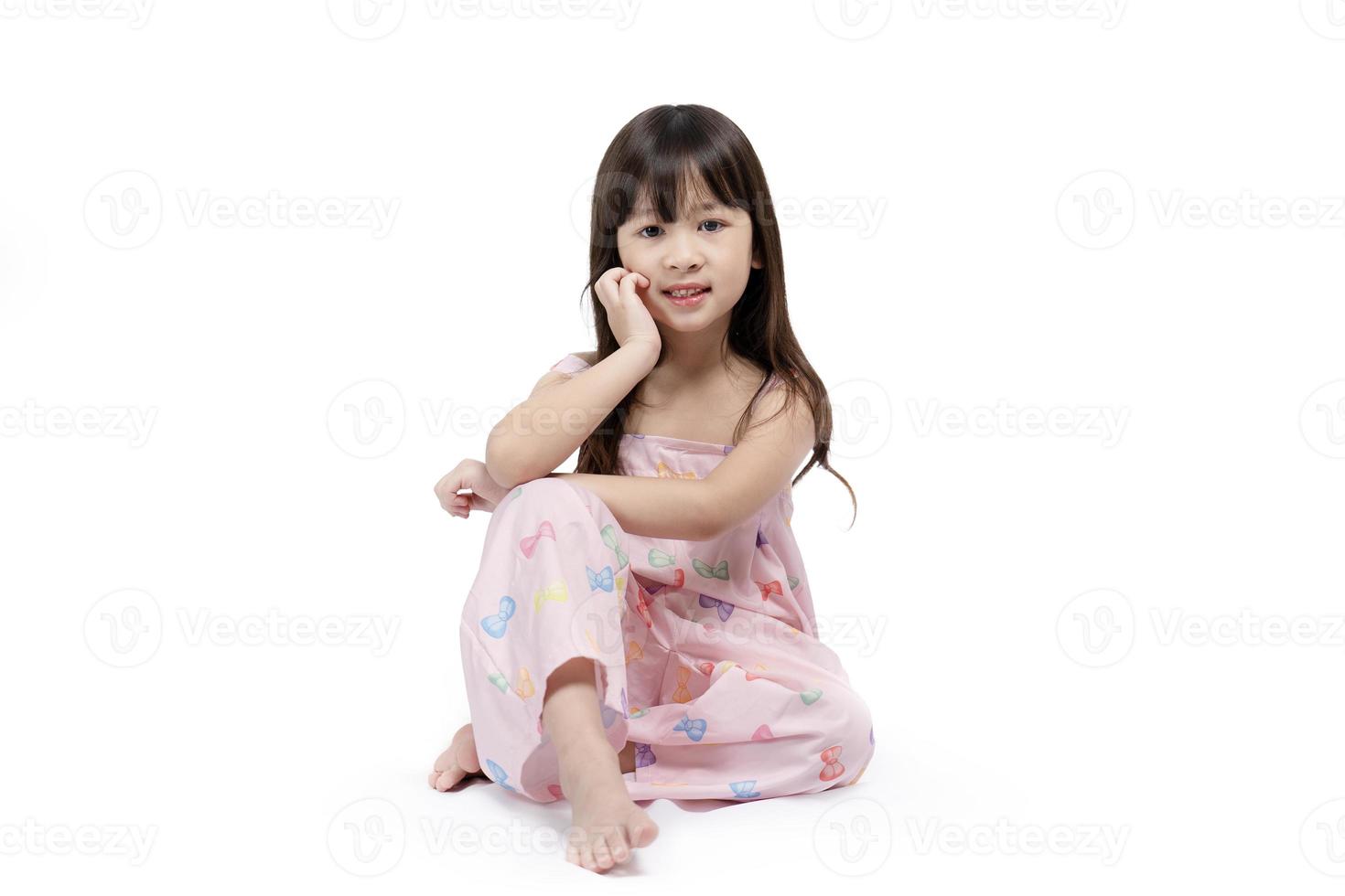 hermosa chica asiática sonriendo brillantemente en un vestido rosa se sienta relajante publicación para sesión de moda sobre fondo blanco. niño lindo feliz, saludable y brillante foto