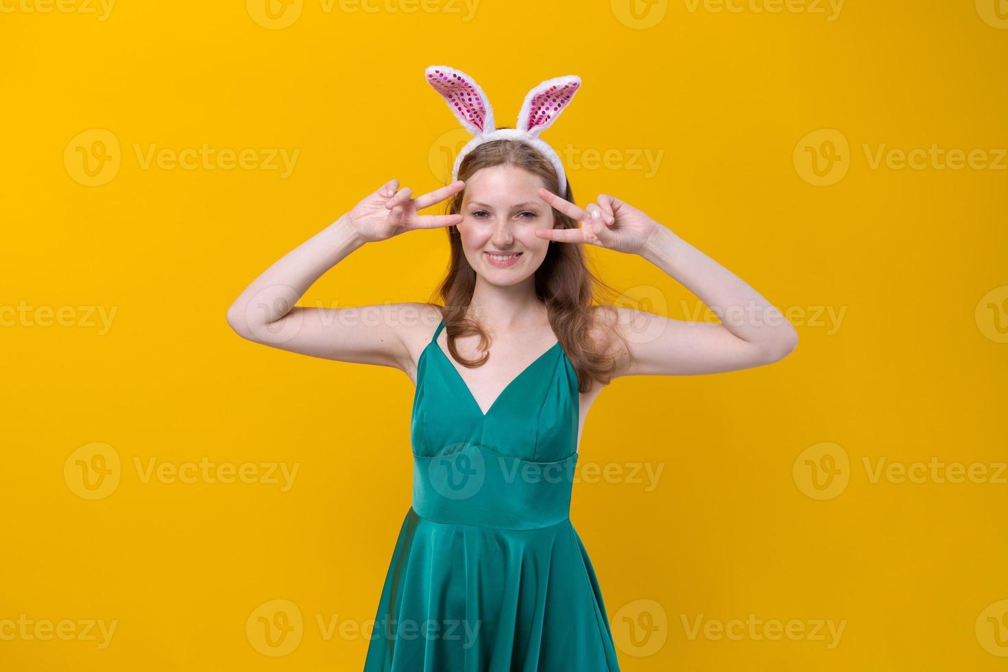 Positive cute girl posing with bunny ears and in green dress on yellow photo