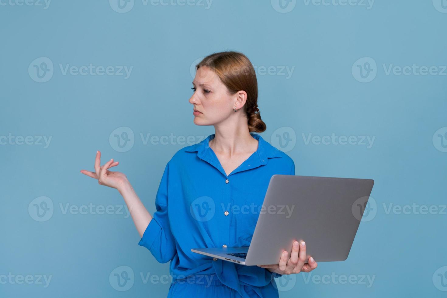Una joven atractiva y seria con una camisa azul lisa parada detrás de una laptop foto
