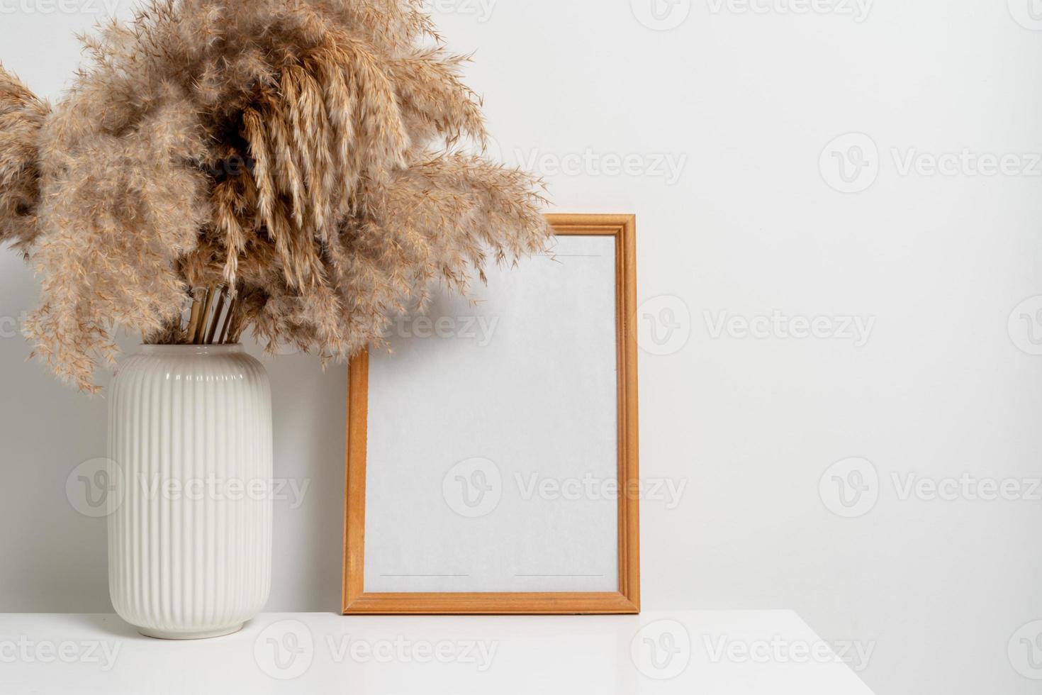 Wooden vertical frame with white vase of dry flowers over white wall photo