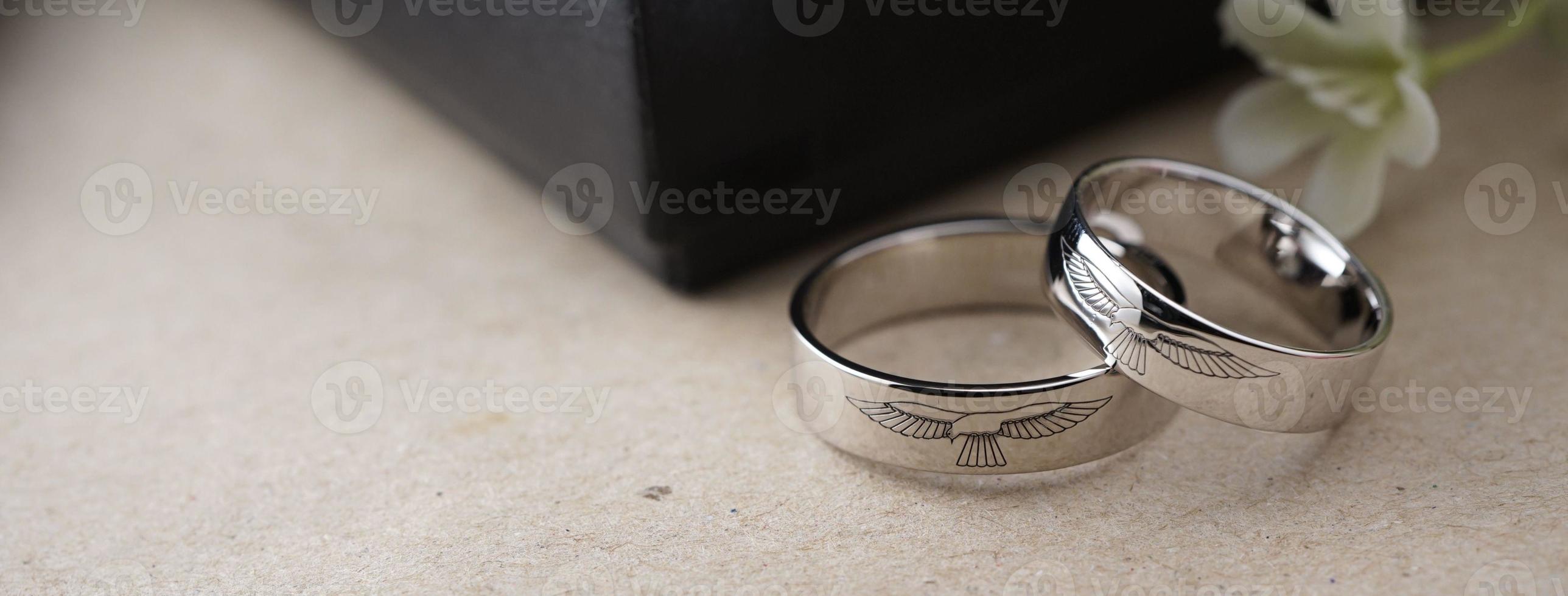 White gold wedding ring with a bird laser on the ring surface. With a black ring box background and artificial flowers. On the left there is an empty space to fill in text photo