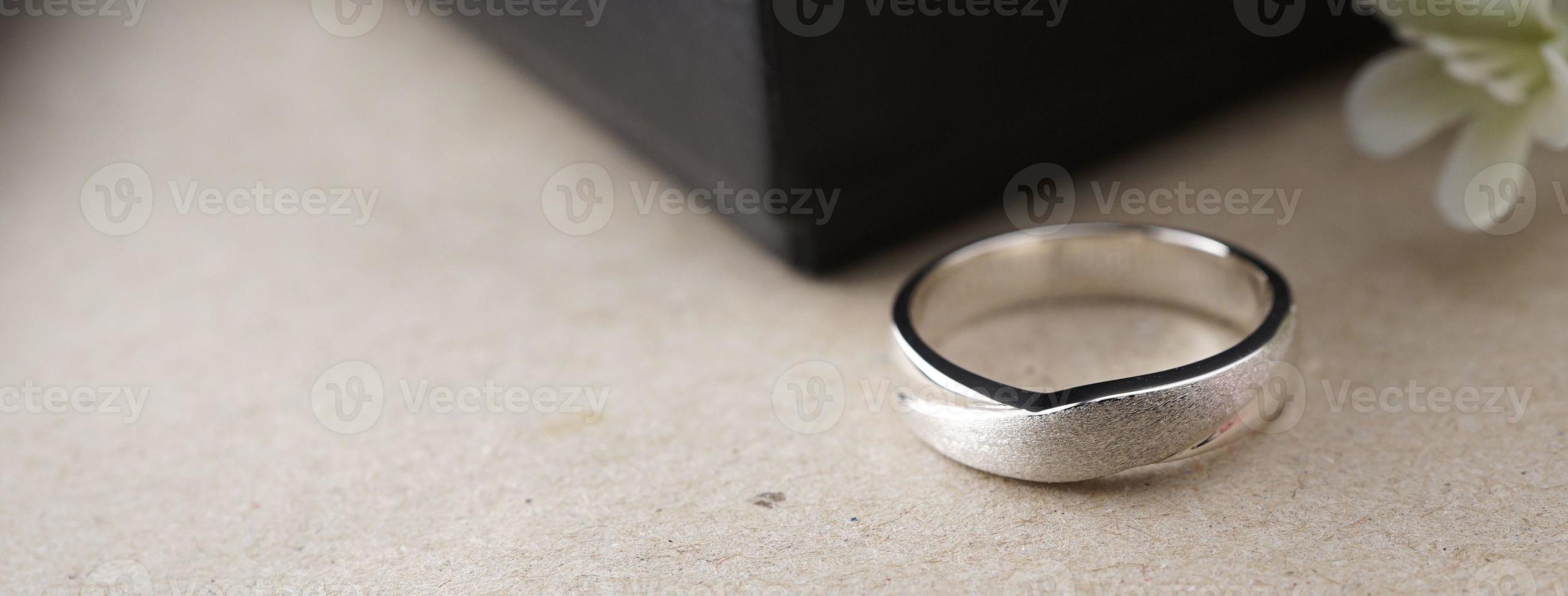 White gold wedding ring with a bird laser on the ring surface. With a black ring box background and artificial flowers. On the left there is an empty space to fill in text photo