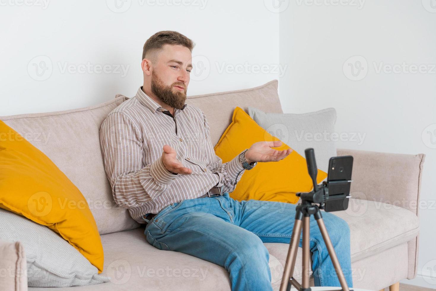 Smiling bearded guy filming video using smartphone on tripod online while photo