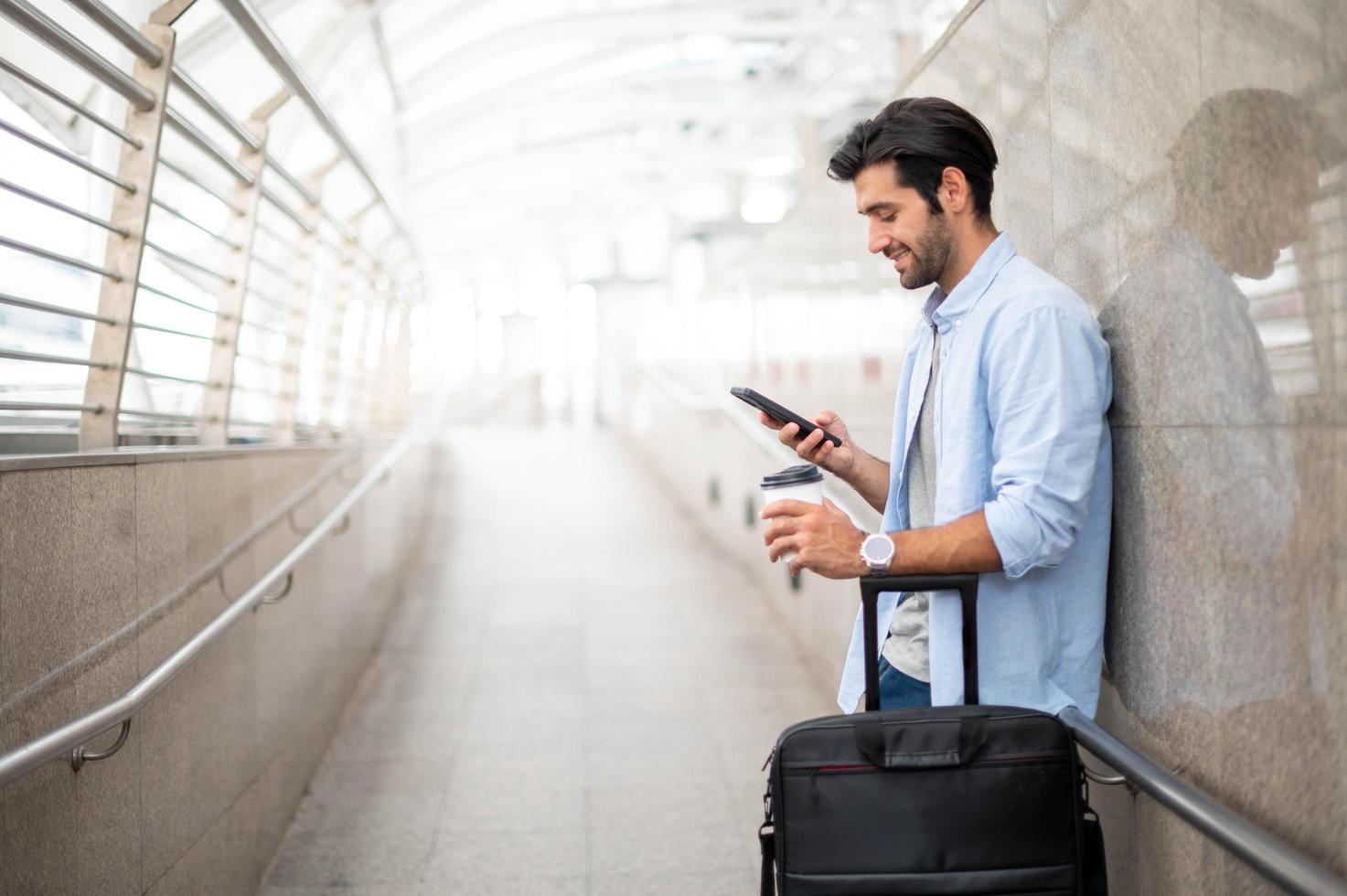 el hombre que usa el teléfono inteligente mientras que la otra mano sostiene una taza de café y tira del equipaje en el aeropuerto. foto