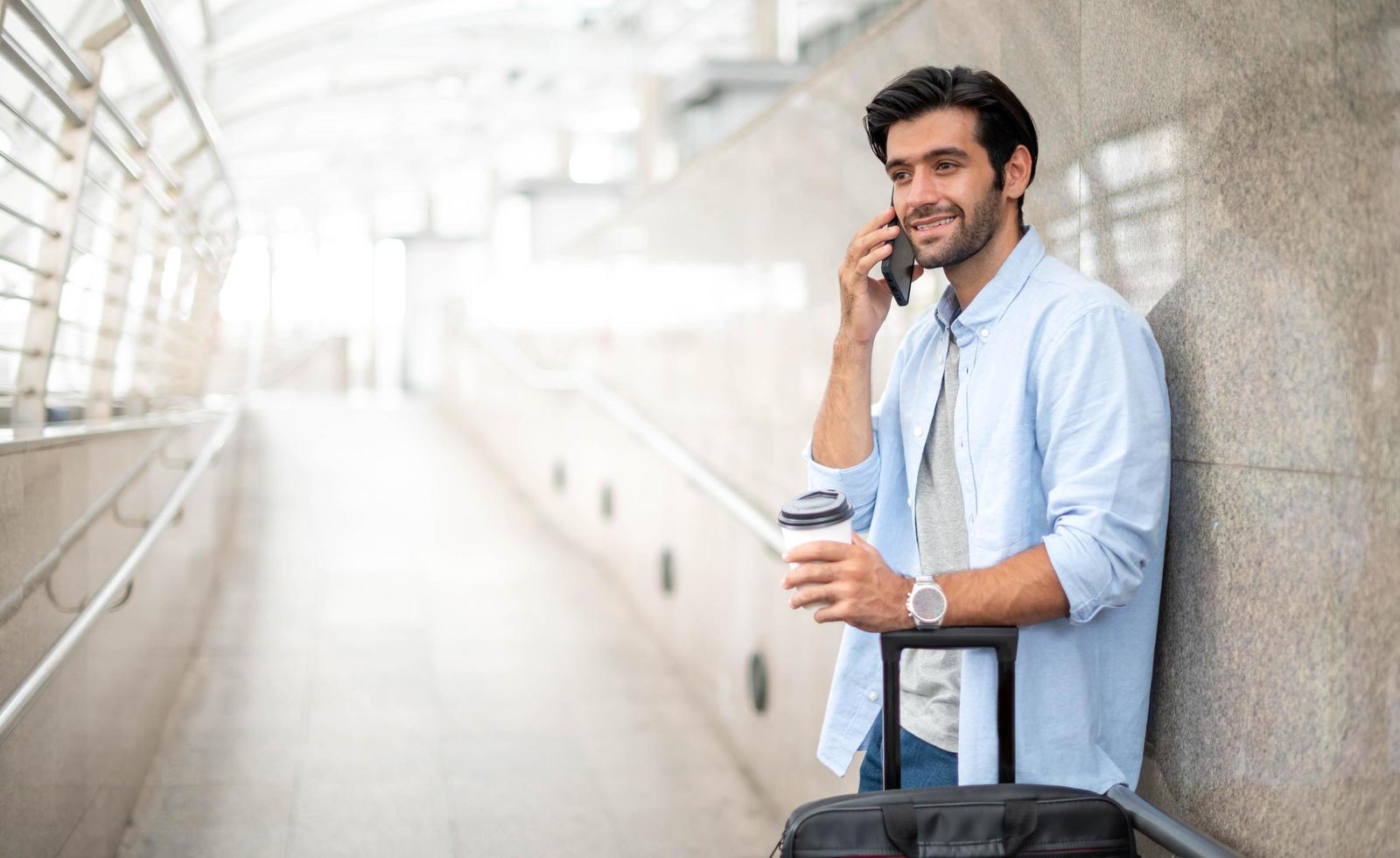 el hombre que usa el teléfono inteligente mientras que la otra mano sostiene una taza de café y tira del equipaje en el aeropuerto. foto