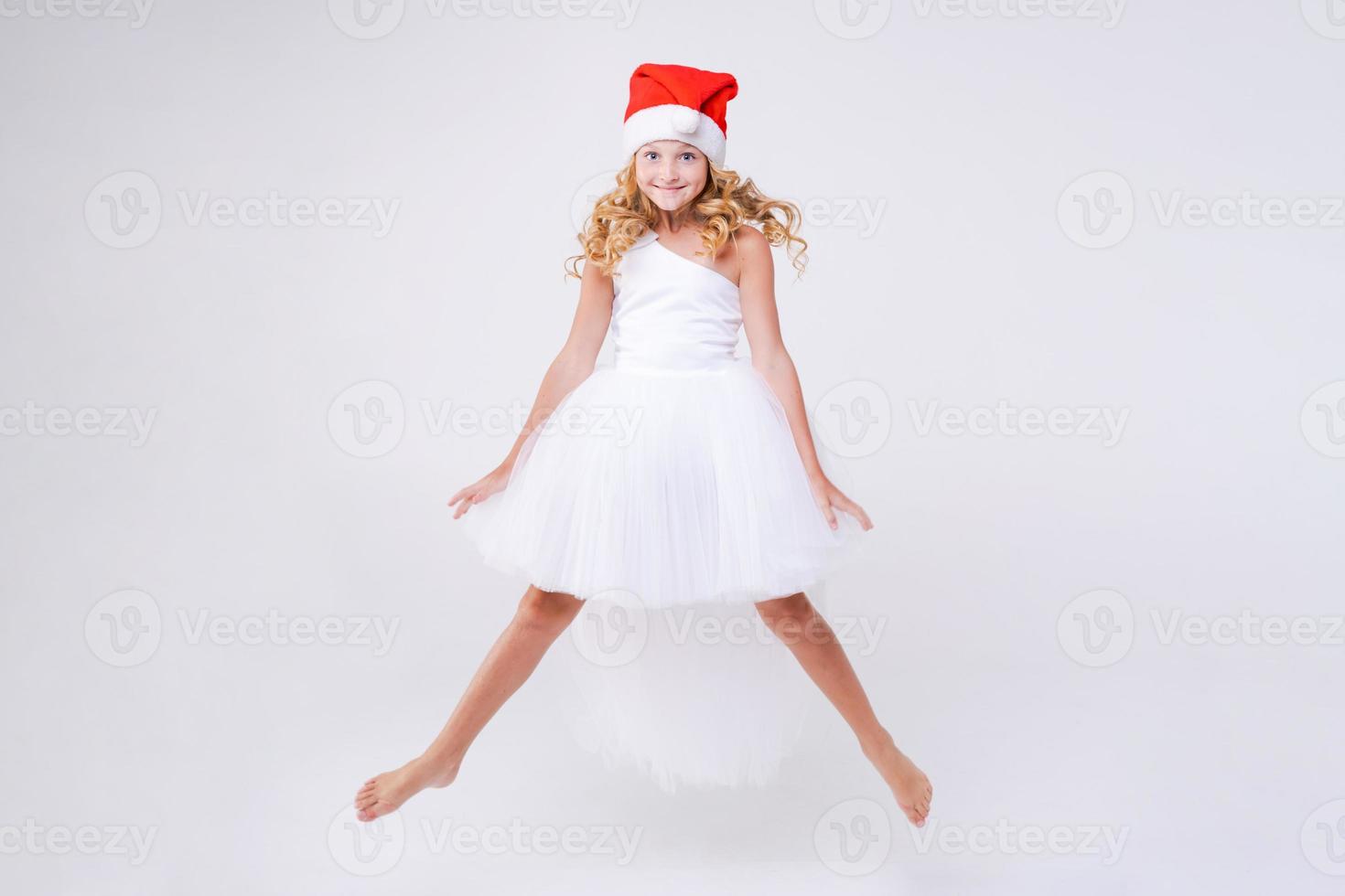 Little joyful girl in red Santa hat and white beautiful dress jumps from photo