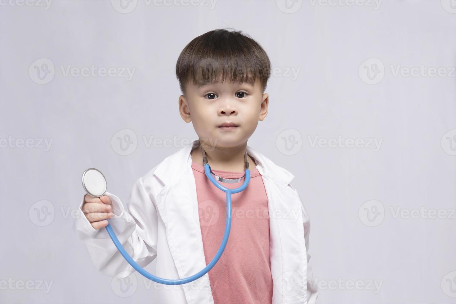 Young cute Asian boy wearing a medical uniform holding a stethoscope playing happy doctor on white background. Preschool children pretend to be a pediatrician. photo