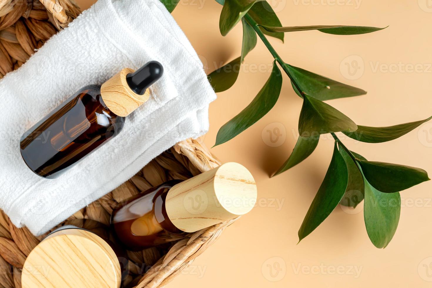 Dark glass and transparent cosmetic bottles with face serum or essentioal oil in a basket with cotton towel on beige background with ruskus leaf. Unbradned Beauty cosmetic products. photo