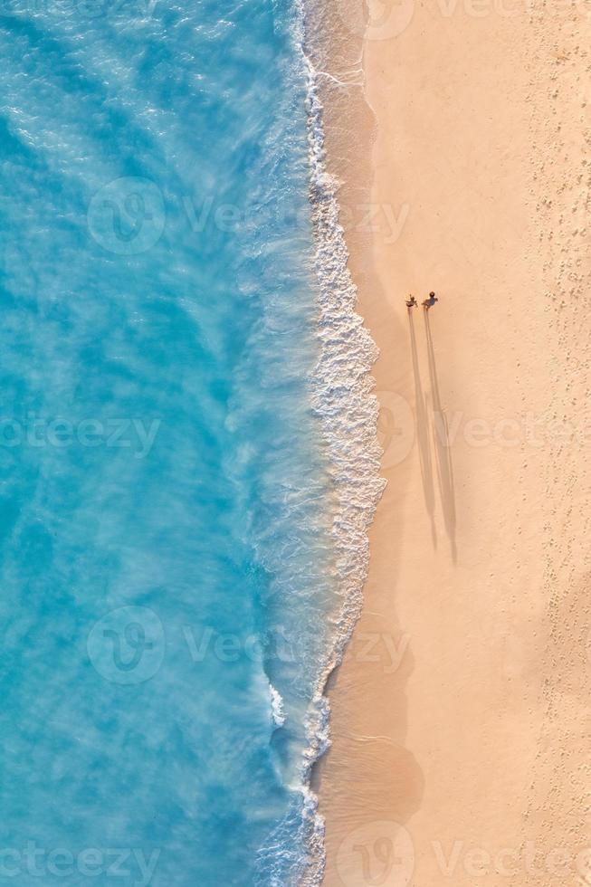 Aerial view of couple walking on beach with sunlight close to turquoise sea waves. Top view of summer beach landscape, romantic inspirational couple vacation, romance holiday. Freedom travel template photo