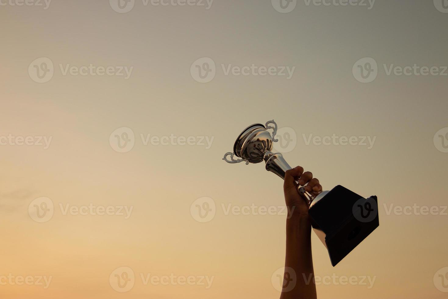 concepto de victoria con una mano sosteniendo un trofeo en el fondo del cielo del atardecer foto