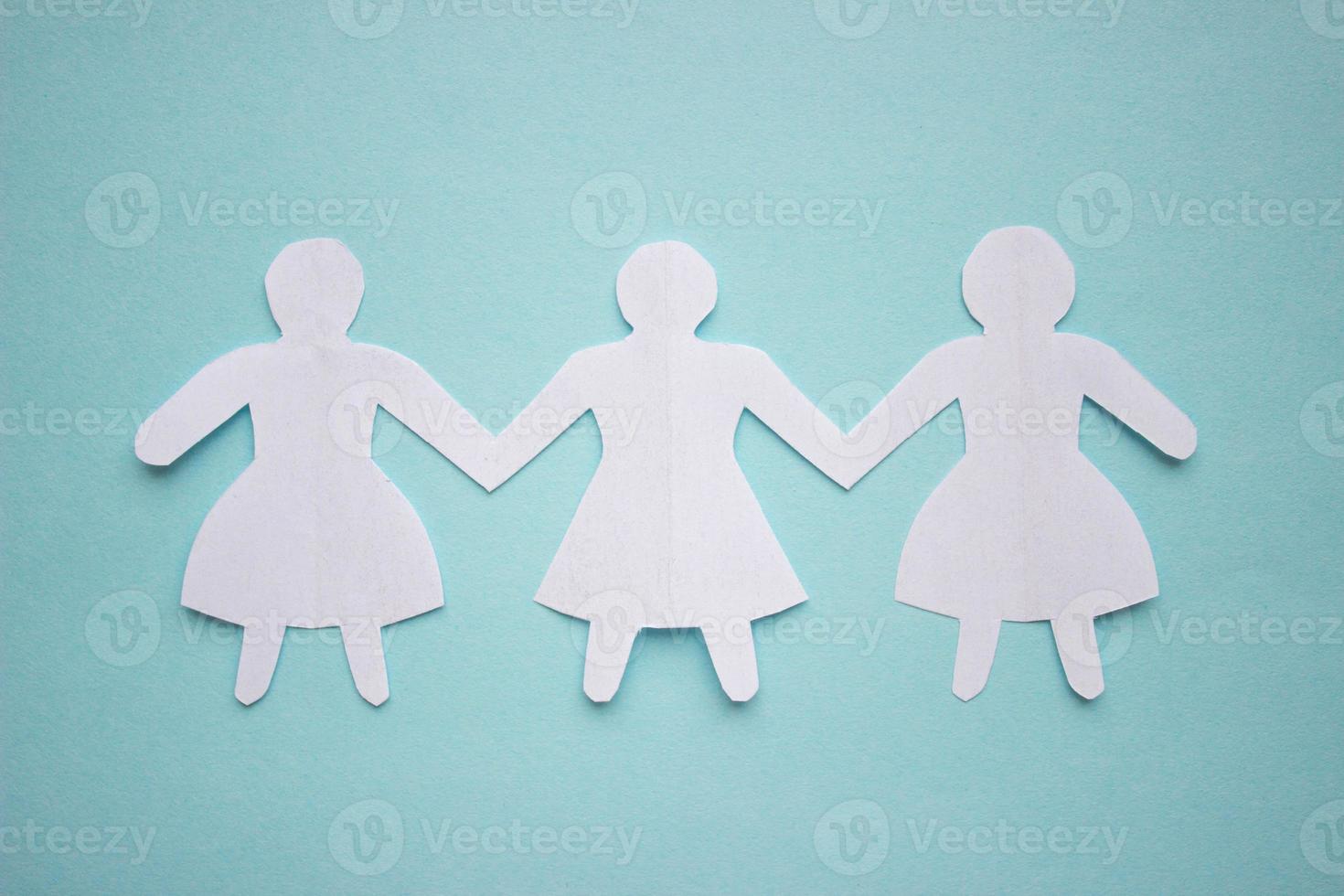 Figures of three girls in dresses holding hands, cut out of white paper. In the center of the photo on a blue background