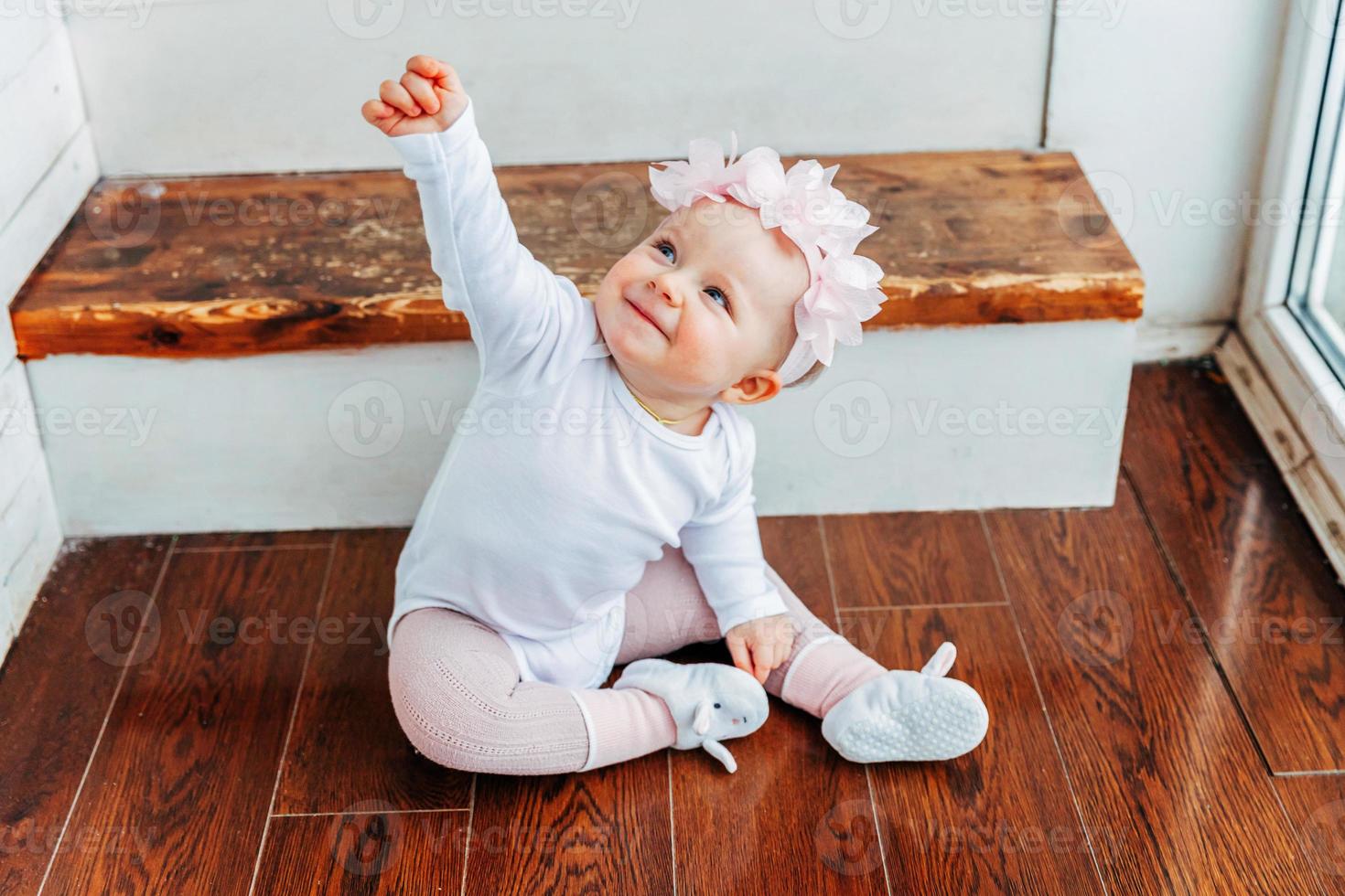 Little smiling baby girl one year old wearing spring wreath siting on floor in bright light living room near window and playing with gerbera flowers. Happy kid playing at home. Childhood concept. photo