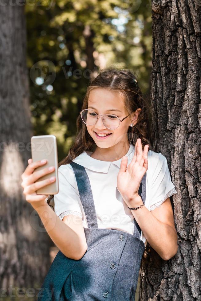 A schoolgirl in glasses makes a video call on a smartphone near a tree in the park. Vertical view photo