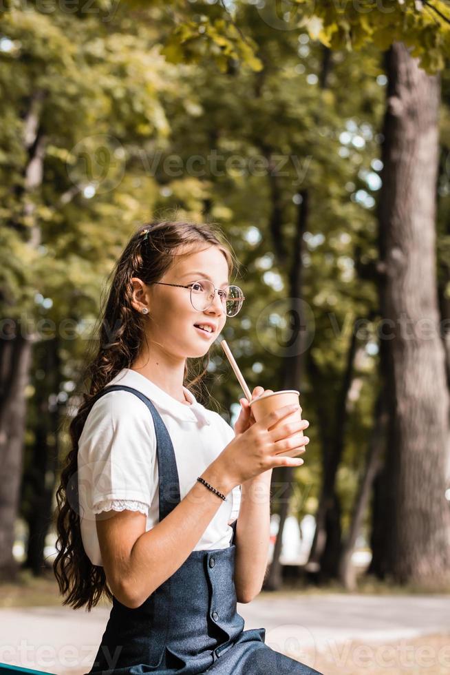 A pupil  in eyeglasses drinks a beverage from an eco paper cup with a straw in the park photo