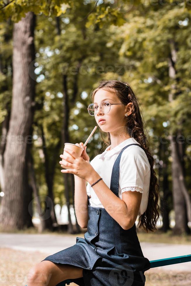 una colegiala con gafas bebe una bebida de un vaso de papel ecológico con una pajita en el parque foto
