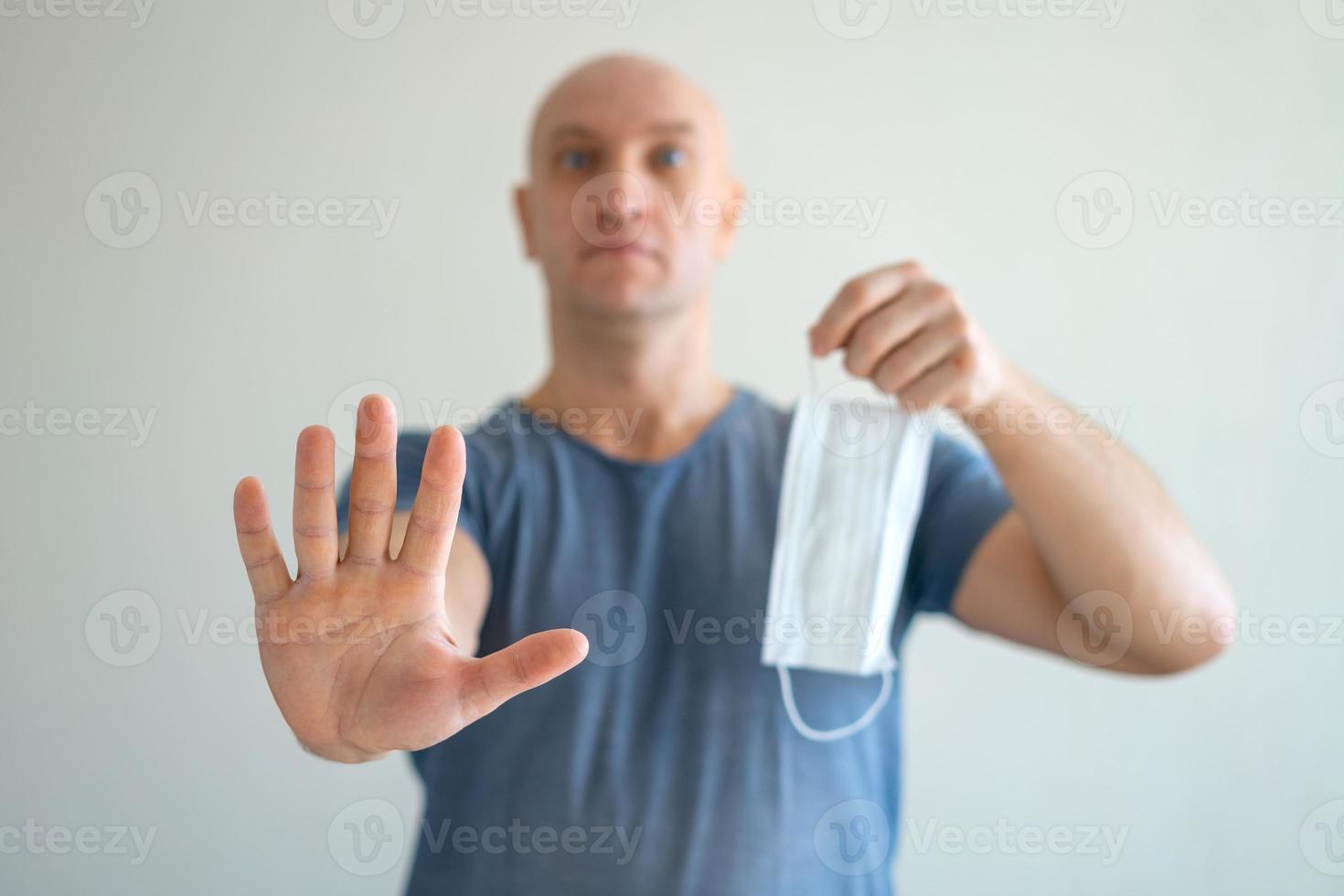 A bald man holds a medical mask in his hands, his other hand held out in protest photo