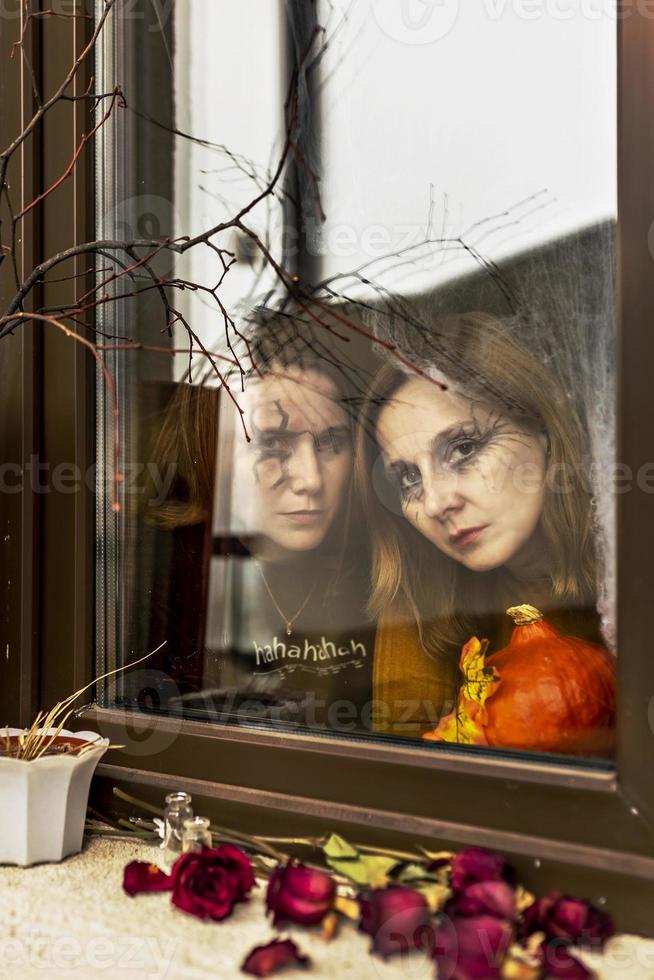 Two female witches look through a spider web ominously out of the window against the backdrop of Halloween decorations. Masquerade, Halloween party photo