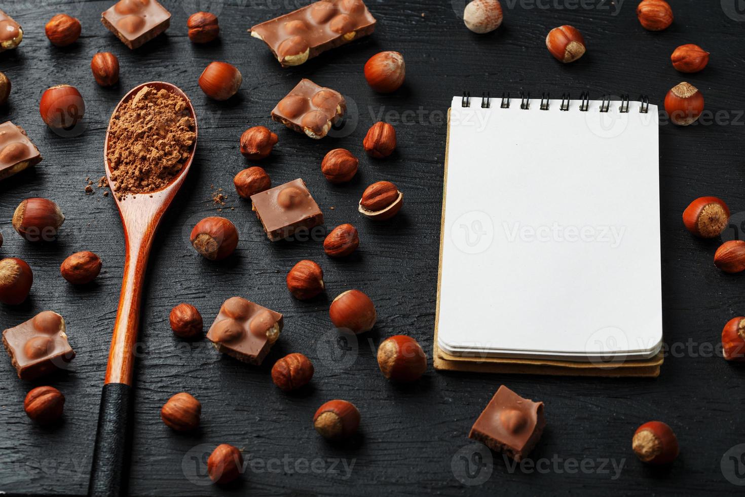 chocolate con avellanas, un cuaderno con páginas en blanco y una cuchara de madera con cacao sobre un fondo oscuro, rodeado de nueces con cáscara y peladas. foto