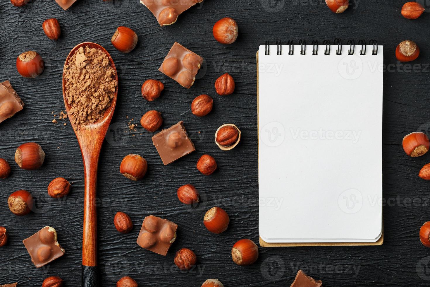 Chocolate with hazelnuts, a notebook with blank pages and a wooden spoon with cocoa on a dark background, surrounded by nuts in the shell and peeled. photo
