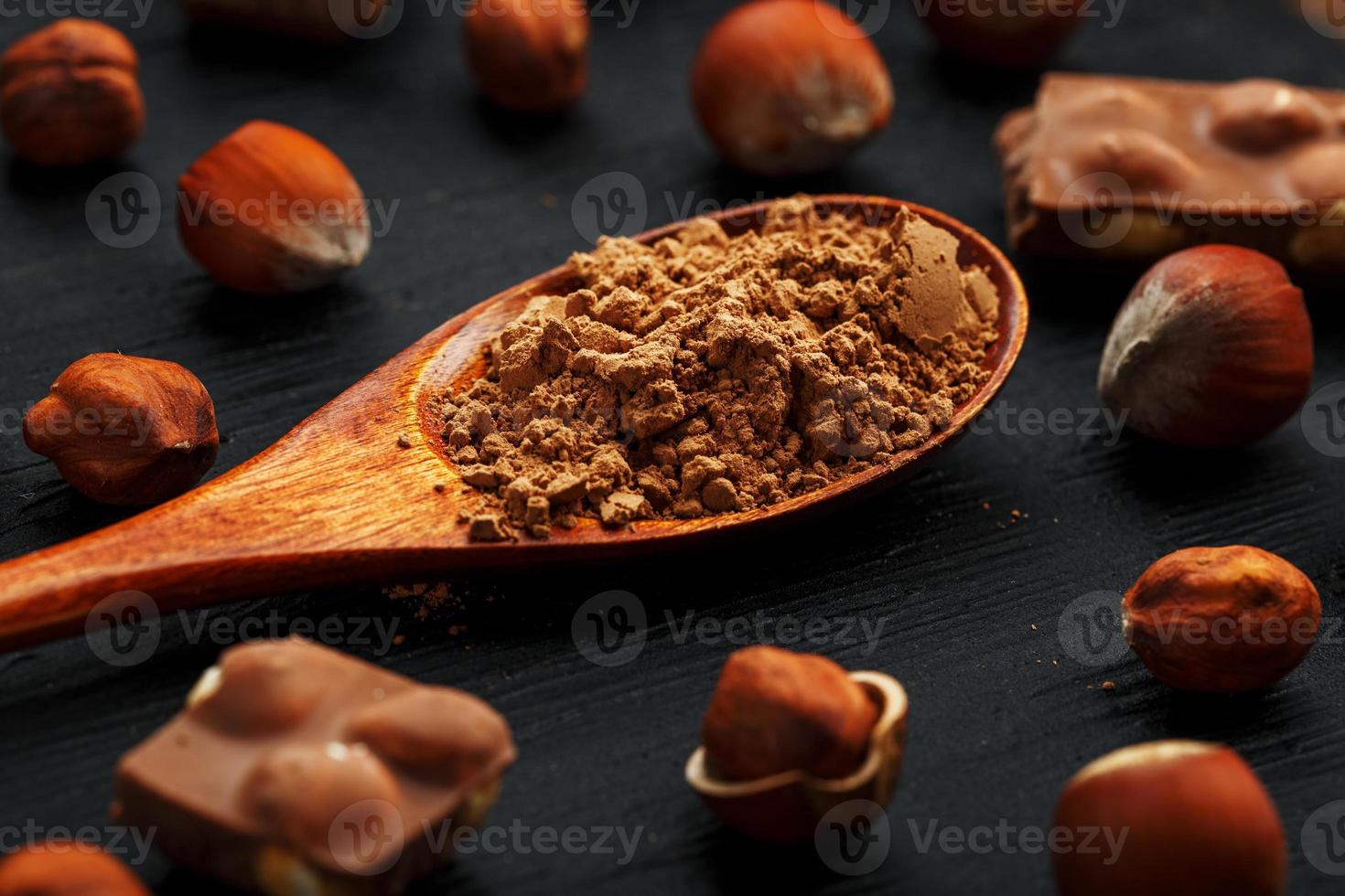 Chocolate with hazelnuts and a wooden spoon with cocoa on a dark background, surrounded by nuts in the shell and peeled. photo