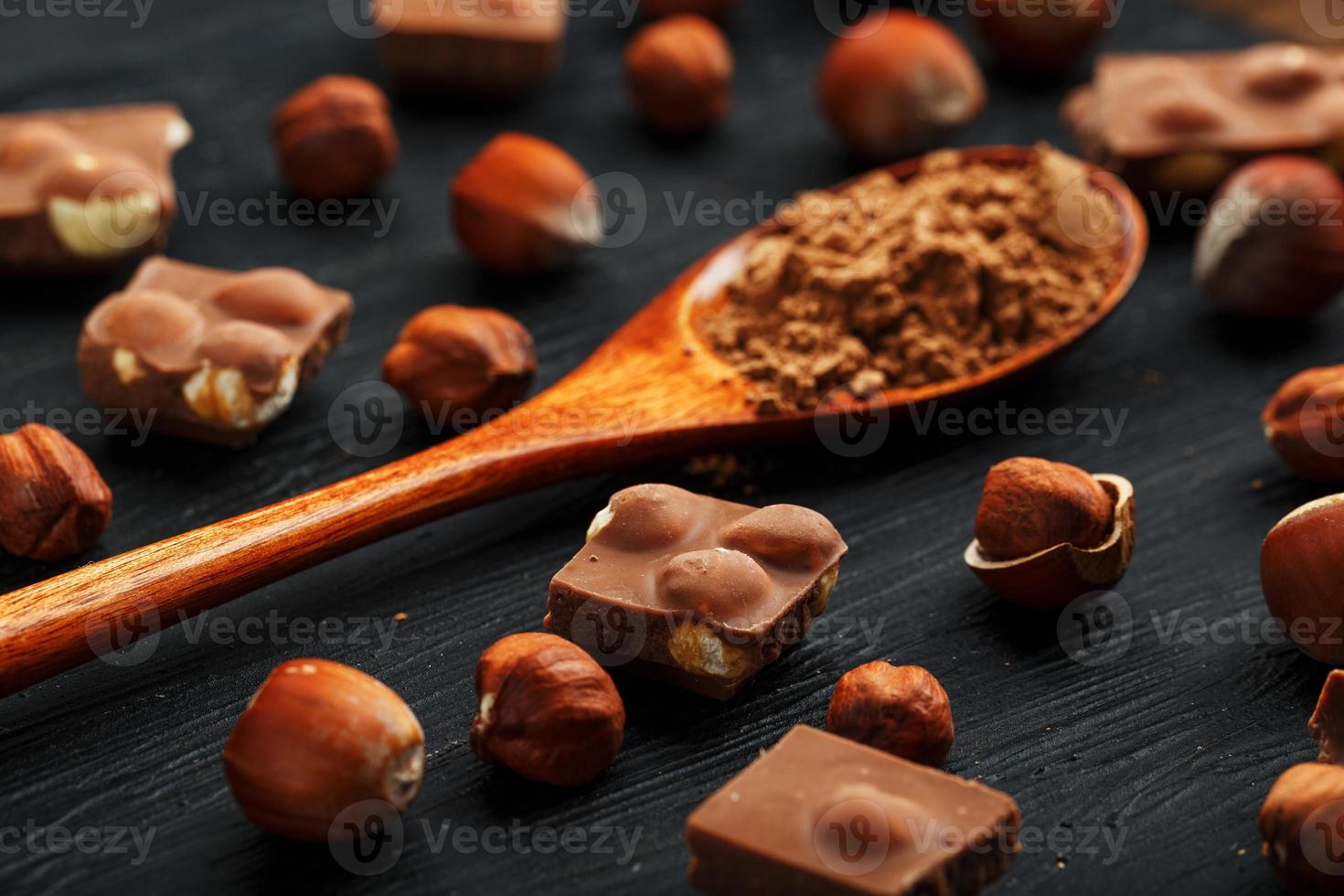 Chocolate with hazelnuts and a wooden spoon with cocoa on a dark background, surrounded by nuts in the shell and peeled. photo