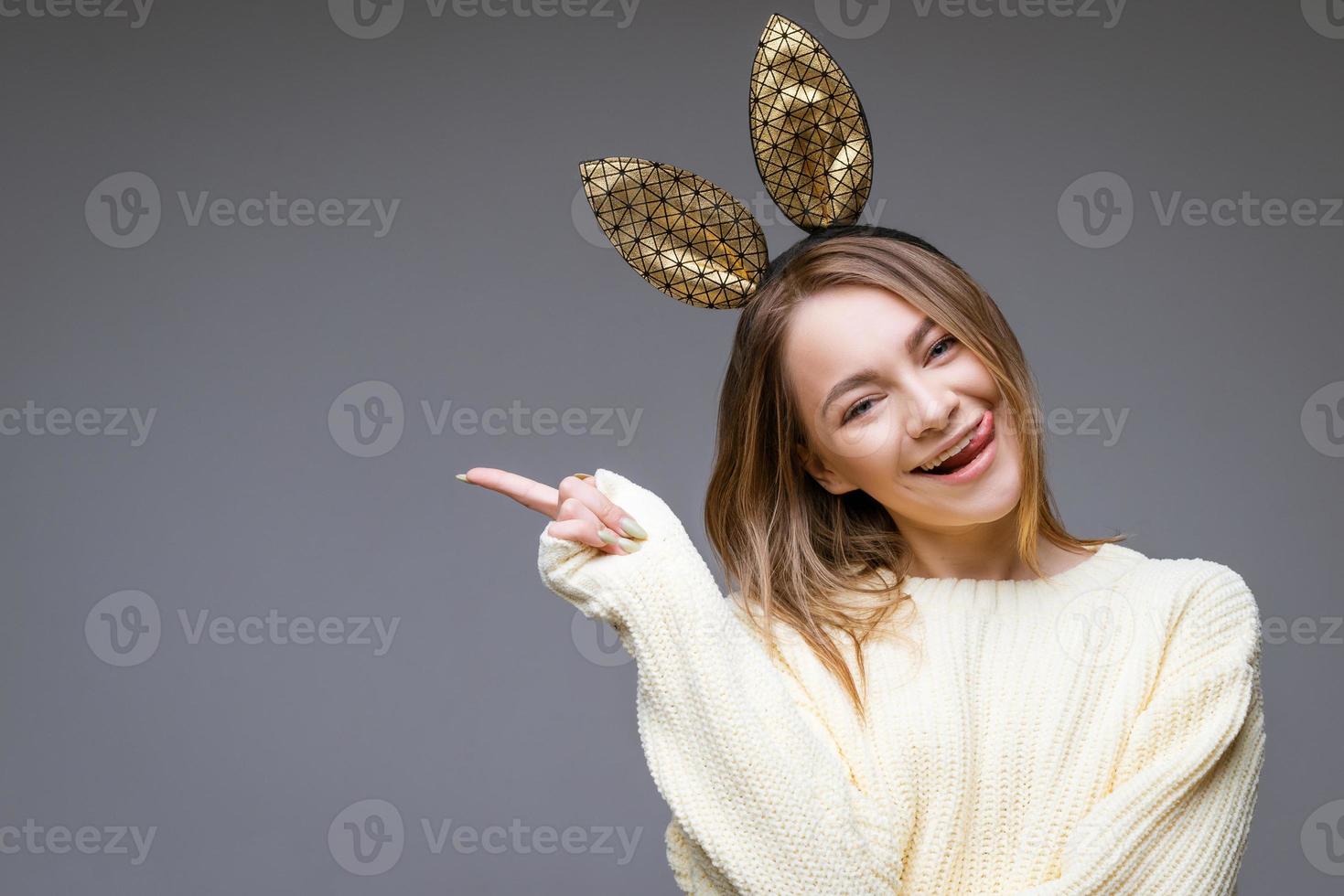 mujer joven en orejas de conejo muestra la lengua y el dedo a un lado en el fondo foto