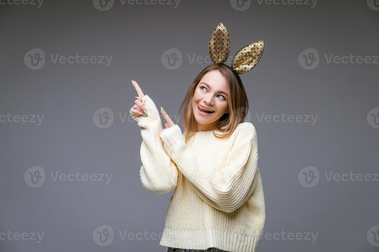 Young woman in rabbit ears shows tongue and finger to the side on background photo