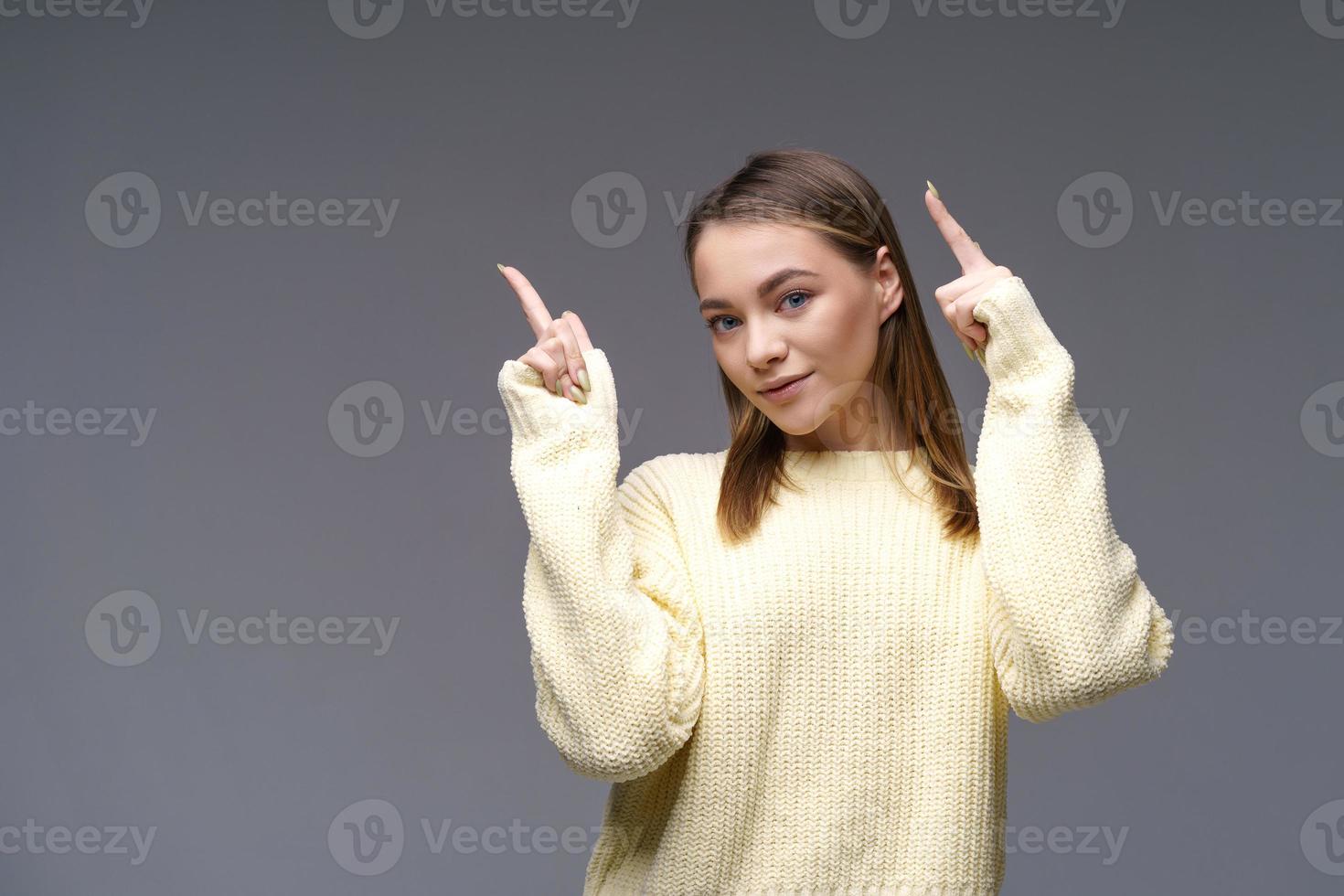 Cheerful young woman showing thumbs up in yellow sweater on gray background photo