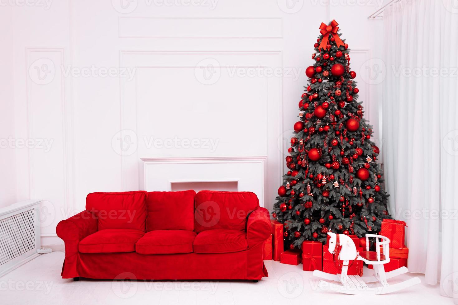 Stylish Christmas minimalist interior with an elegant red sofa. Comfort home with fireplace. Gifts boxes under Christmas tree with decorations red balls in white living room. photo