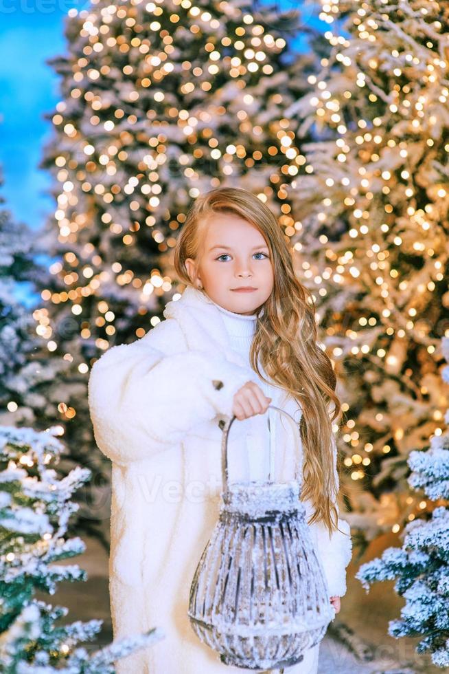cute caucasian blonde girl in white eco fur coat walking in winter christmas forest with lights. New year, fairy tale concept photo