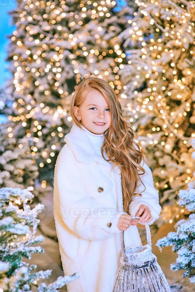 cute caucasian blonde girl in white eco fur coat walking in winter christmas forest with lights. New year, fairy tale concept photo