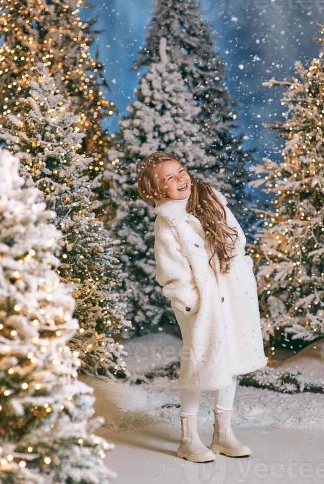 cute caucasian blonde girl in white eco fur coat walking in winter christmas forest with lights. New year, fairy tale concept photo