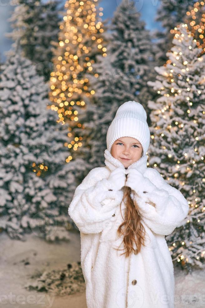Linda chica rubia caucásica en abrigo de piel ecológica blanca, sombrero y guantes caminando en el bosque de Navidad de invierno. año nuevo, cuento de hadas, concepto de moda. foto