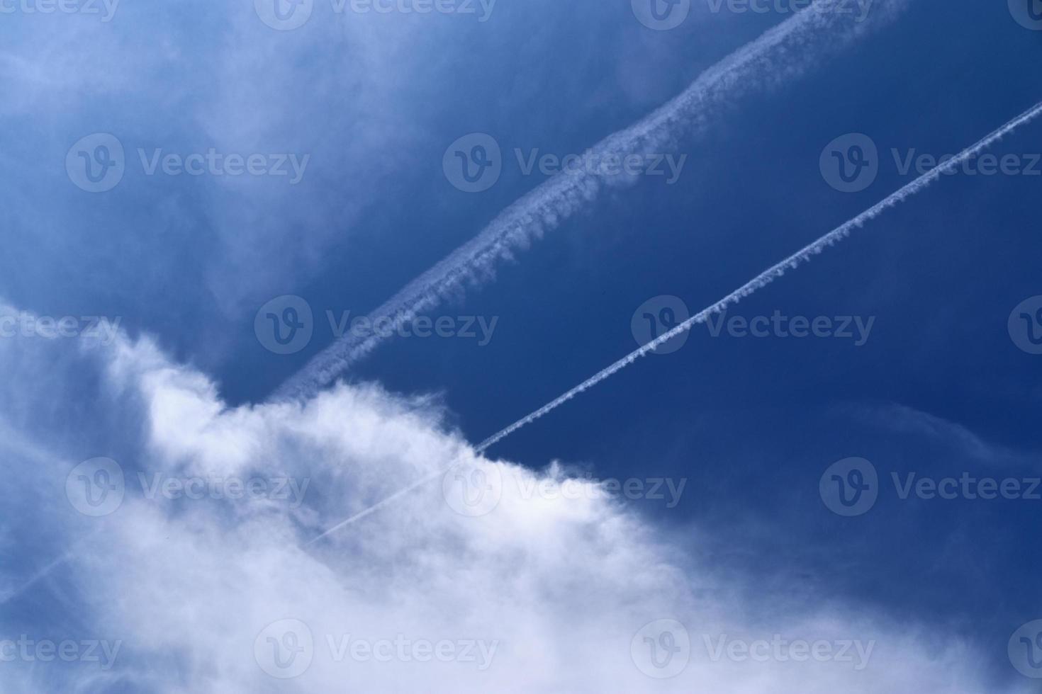 Aircraft condensation contrails in the blue sky inbetween some clouds photo