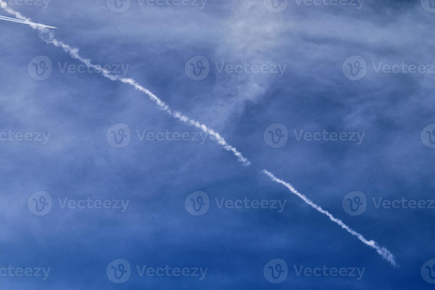 Aircraft condensation contrails in the blue sky inbetween some clouds photo