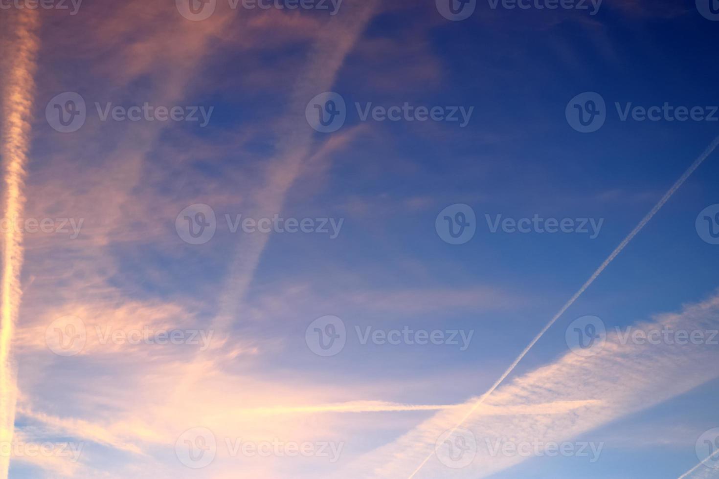 Aircraft condensation contrails in the blue sky inbetween some clouds photo