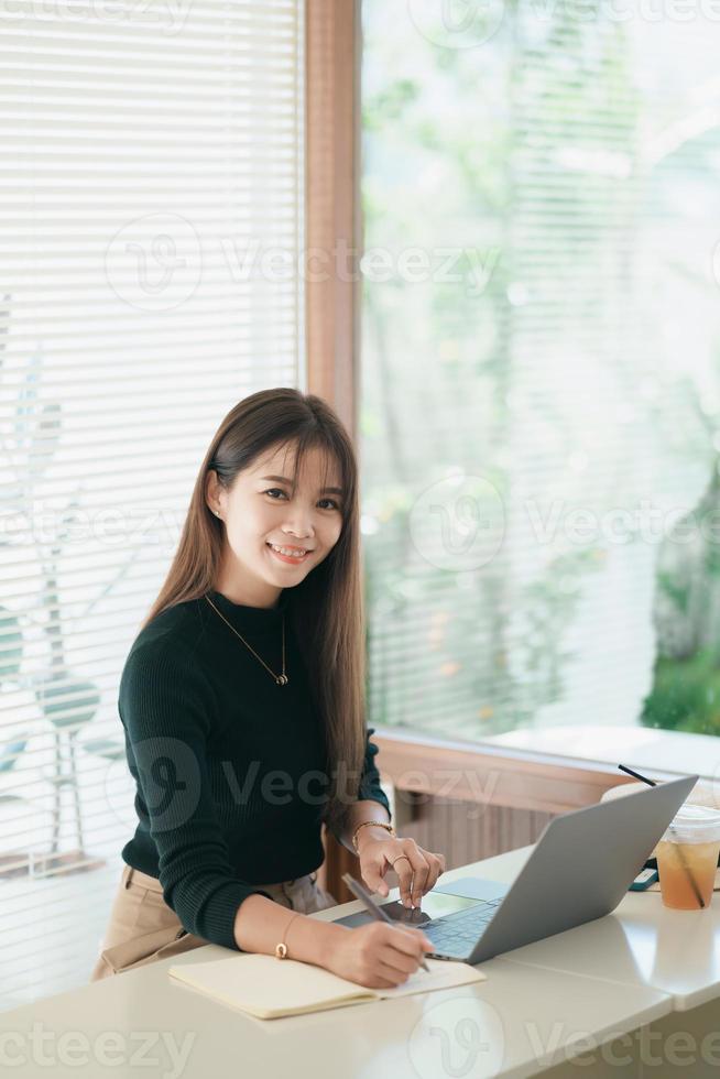 Asian freelance woman smile typing on keyboard and working on laptop on wooden table at home. Entrepreneur woman working for her business at the cafe. Business work at anywhere concept. photo