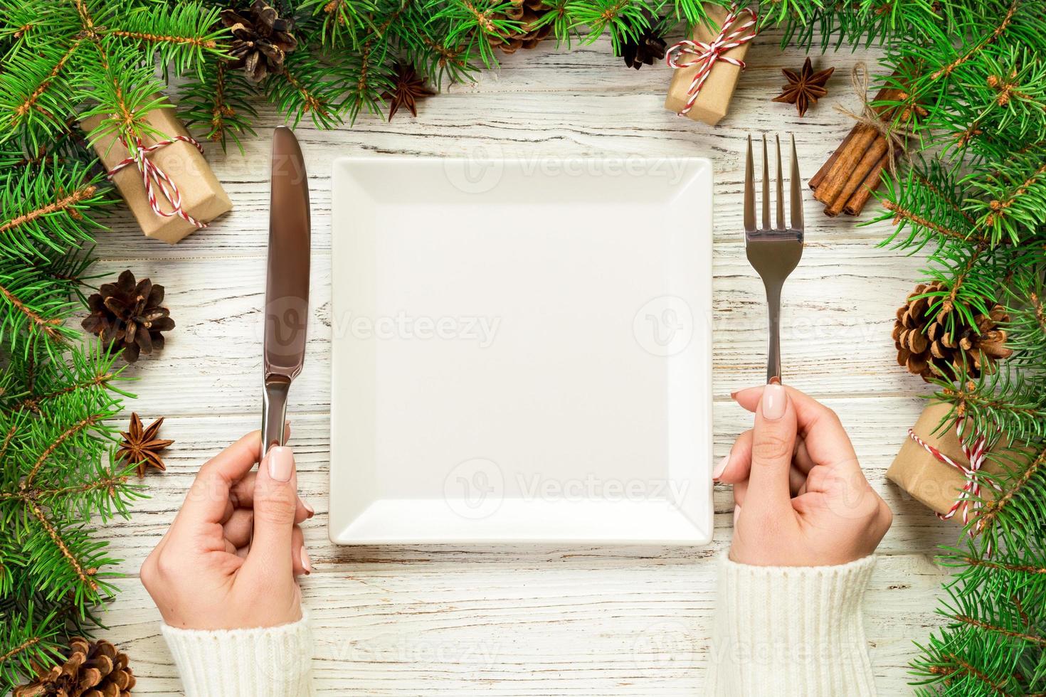 top view girl holds fork and knife in hand and is ready to eat. Empty white square plate on wooden christmas background. holiday dinner dish concept with new year decor photo