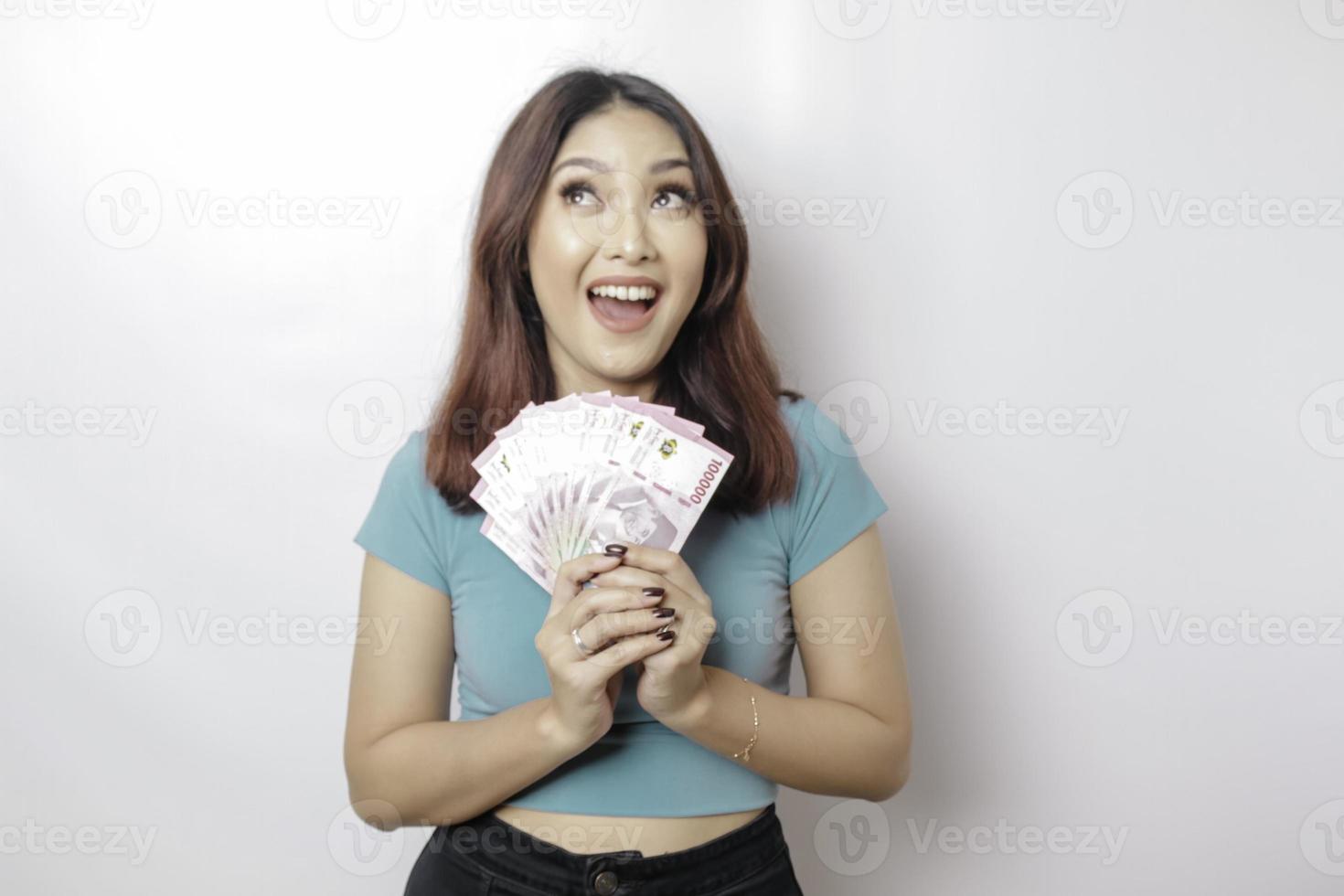 una joven feliz lleva una camiseta azul y tiene dinero en efectivo en rupias indonesias aislada de fondo blanco foto