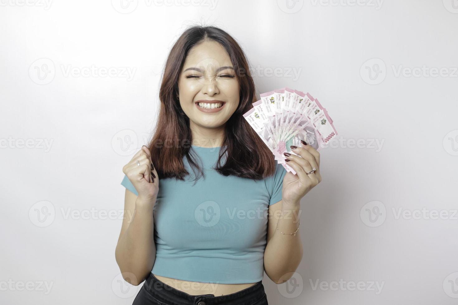 una joven feliz lleva una camiseta azul y tiene dinero en efectivo en rupias indonesias aislada de fondo blanco foto