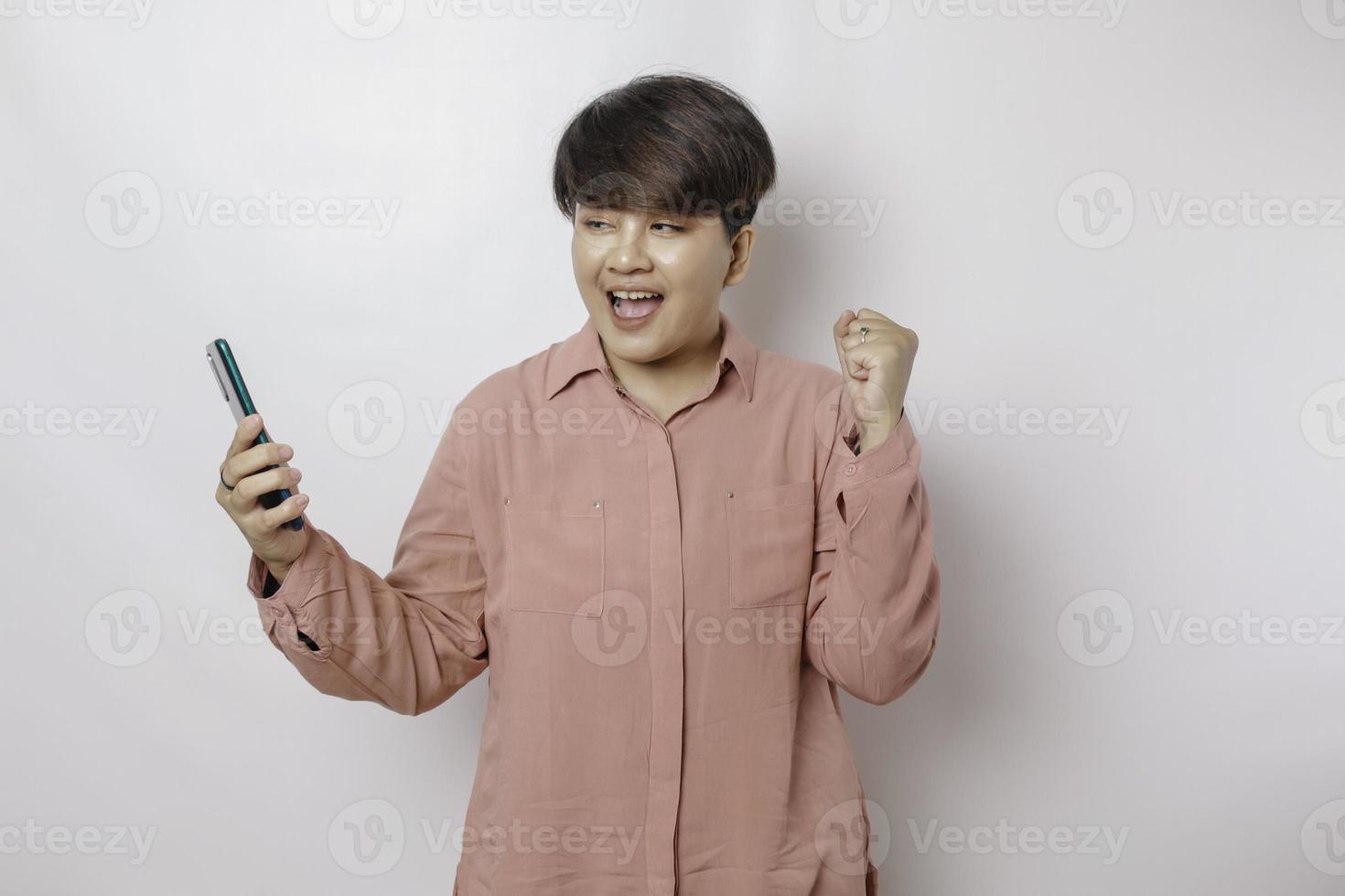 A portrait of a happy Asian woman is smiling and holding her smartphone wearing a pink shirt isolated by a white background photo