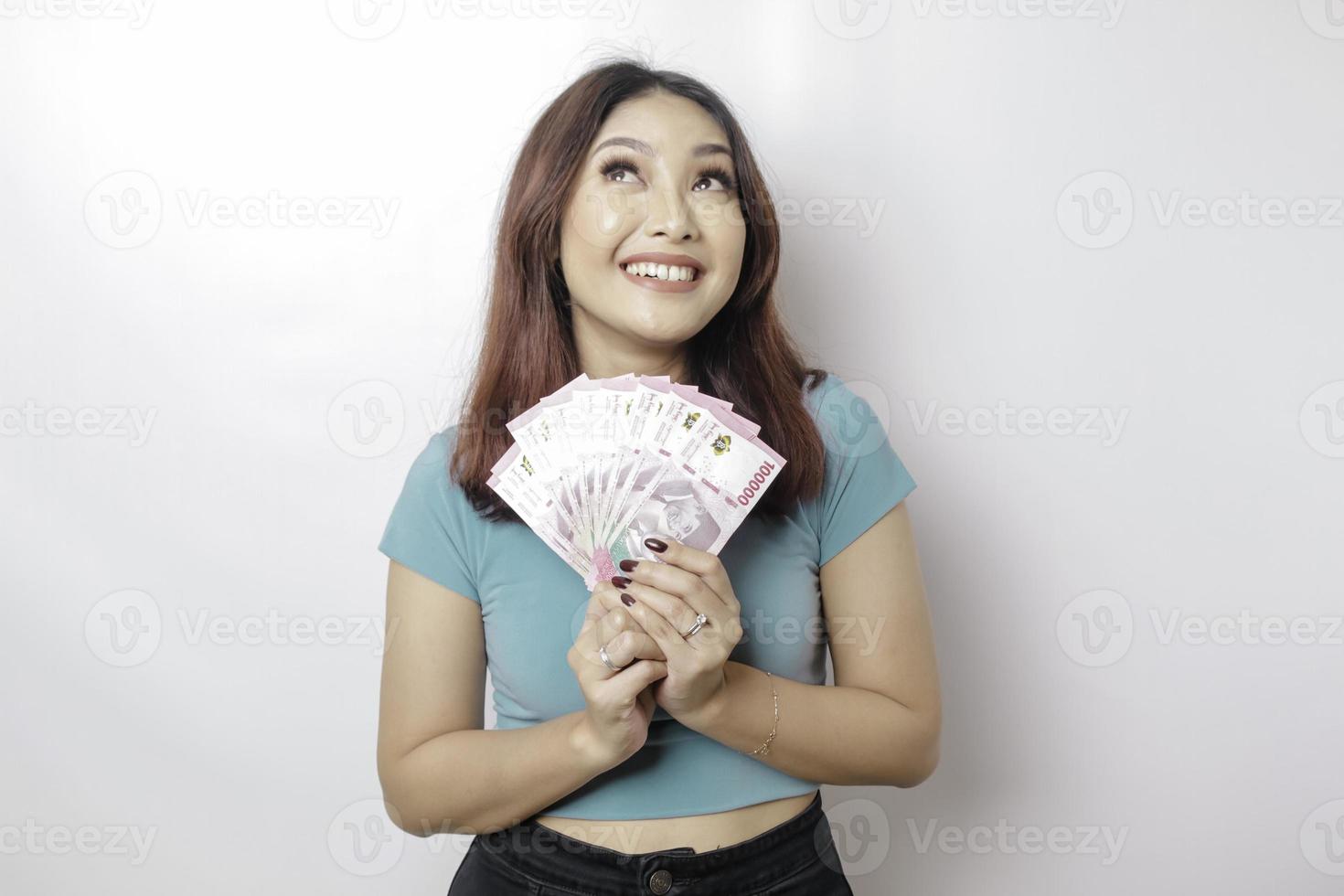 una joven feliz lleva una camiseta azul y tiene dinero en efectivo en rupias indonesias aislada de fondo blanco foto