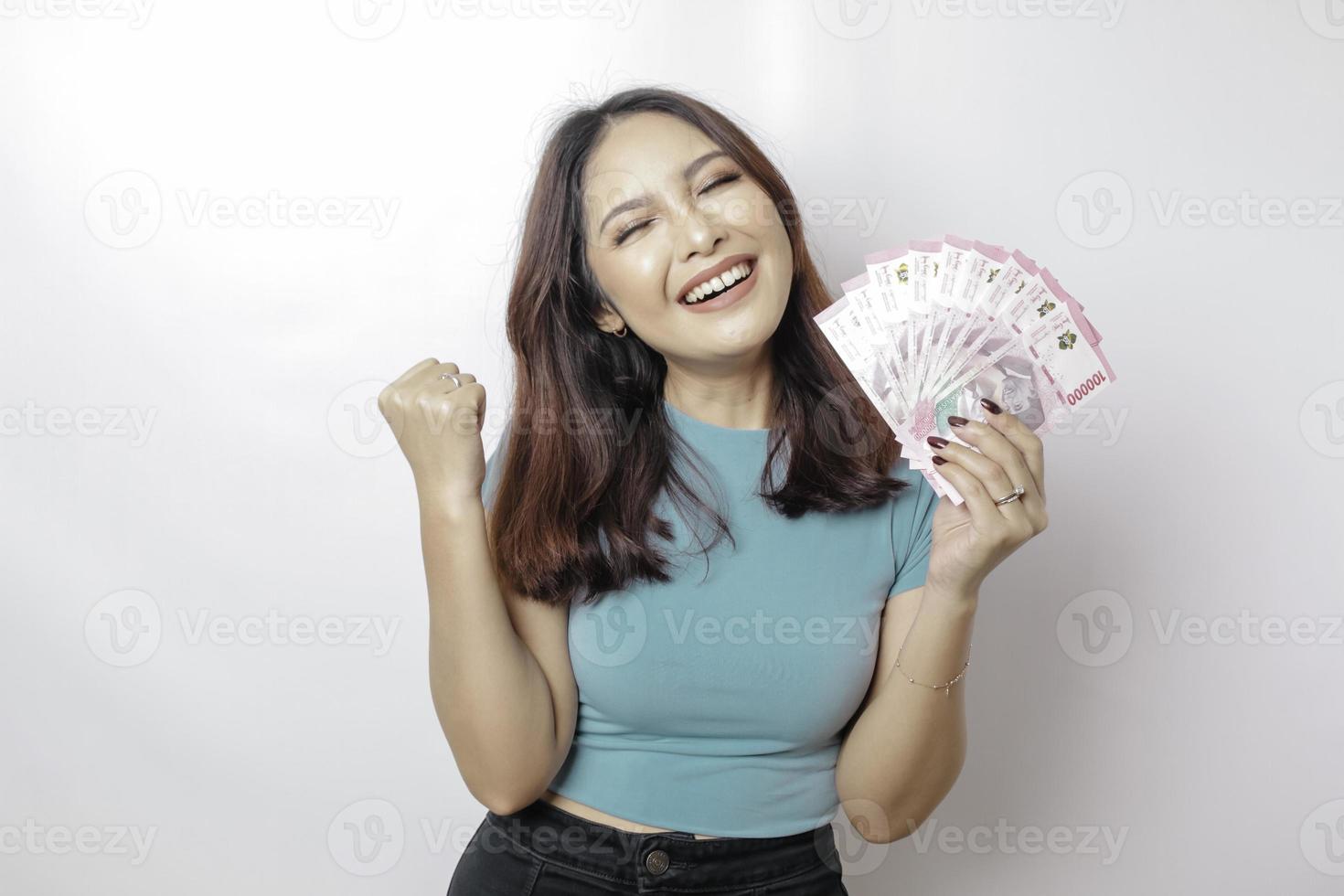 una joven feliz lleva una camiseta azul y tiene dinero en efectivo en rupias indonesias aislada de fondo blanco foto