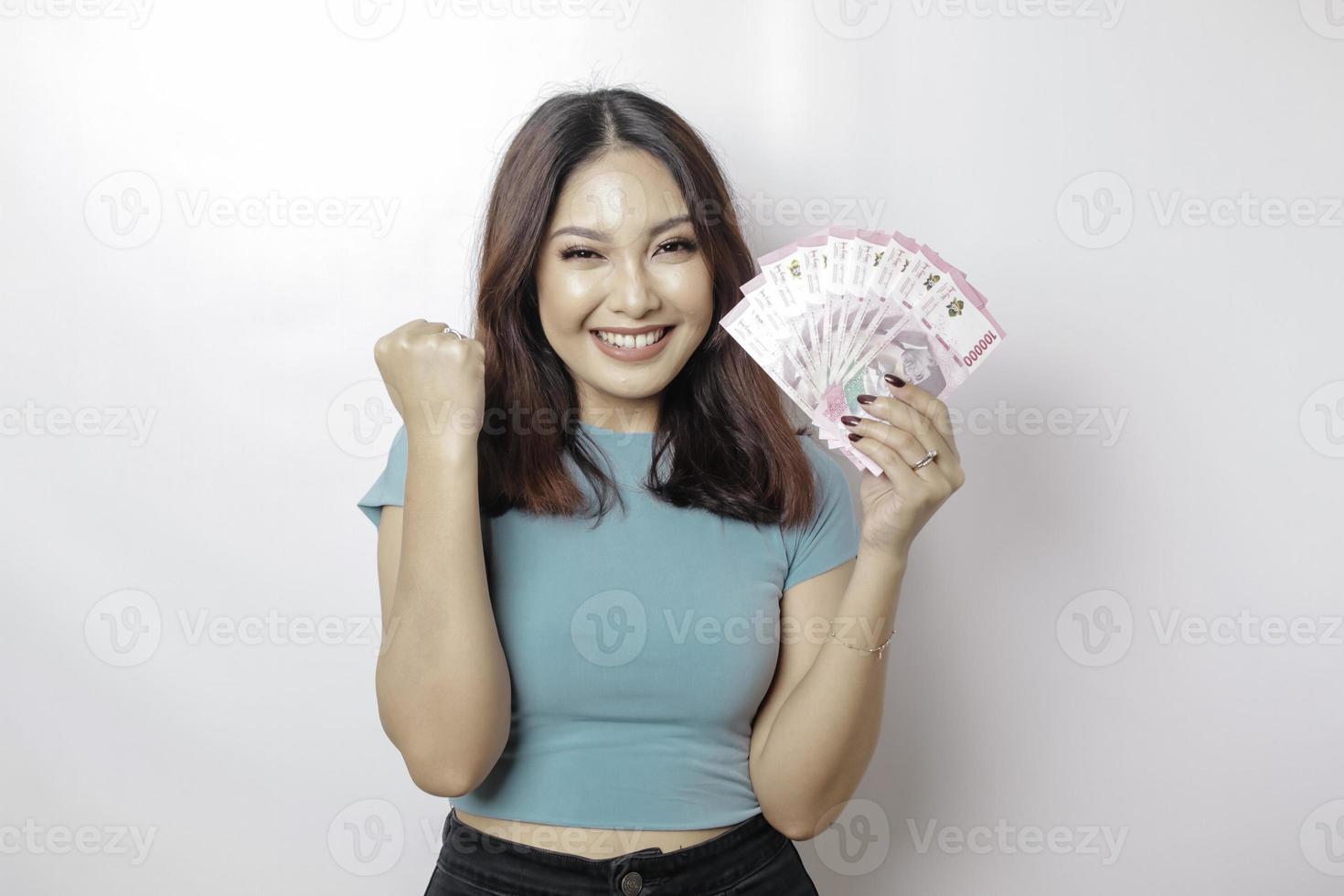 una joven feliz lleva una camiseta azul y tiene dinero en efectivo en rupias indonesias aislada de fondo blanco foto