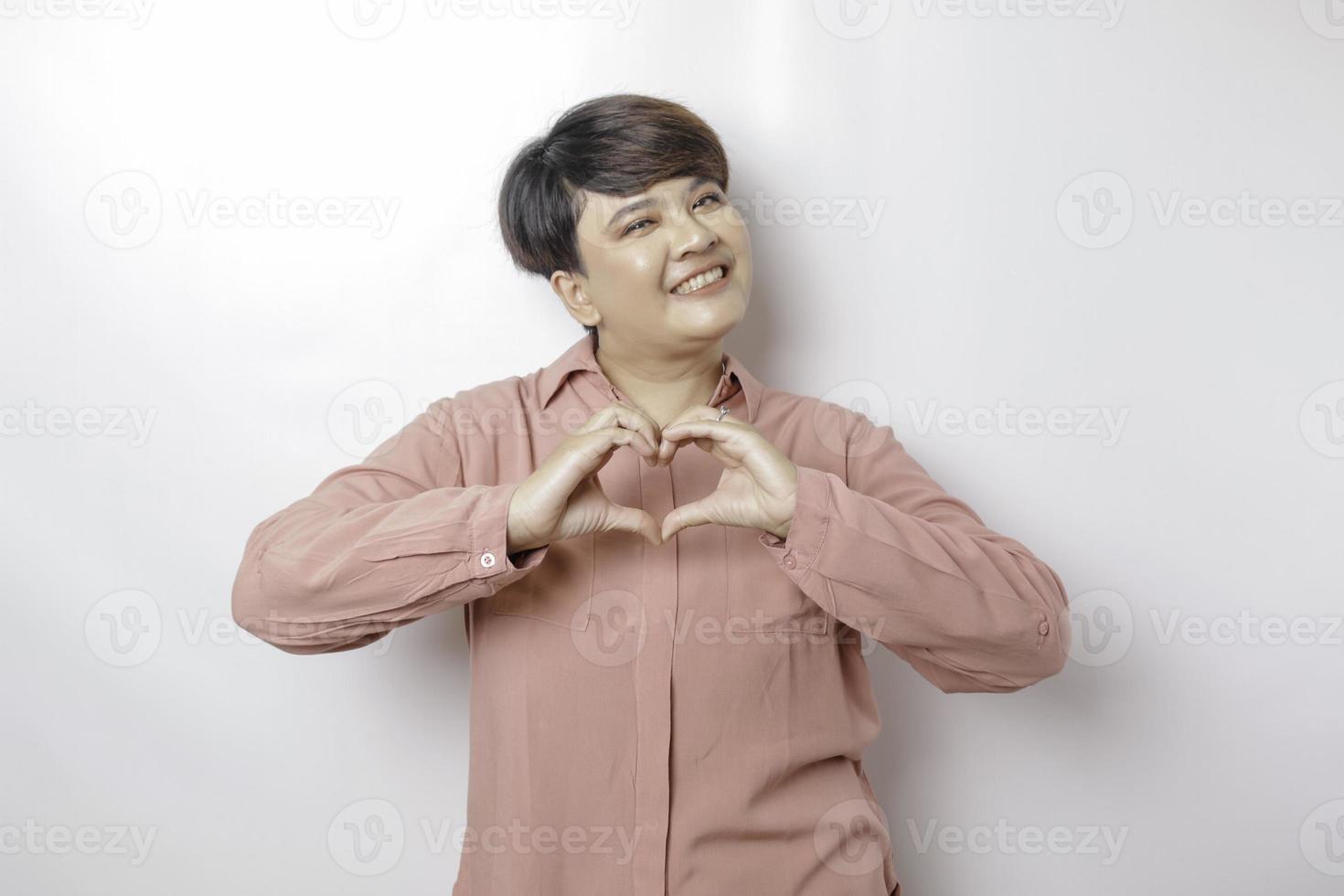 An attractive young short-haired Asian woman wearing a pink shirt feels happy and a romantic shapes heart gesture expresses tender feelings photo